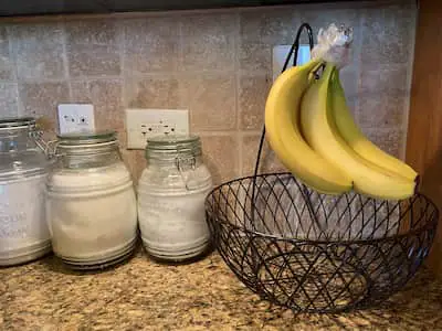 Bananas with stems wrapped in plastic hanging from an empty fruit bowl.