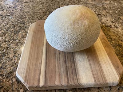 1 whole cantaloupe on a wooden cutting board on a granite counter top.
