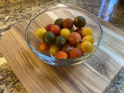 Cherry heirloom tomatoes in a glass bowl on a wooden cutting board on a granite counter top.
